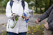 Oben: Eine Person mit weißer Jacke und blauem Rücksack zeigt gesammelte Glasflaschen und Plastik. Die Person rechts von der ist kaum zu sehen gibt aber ein "double thumbs up"